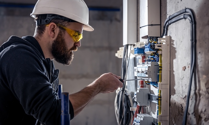 A male electrician works in a switchboard with an electrical connecting cable, connects the equipment with tools.